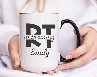 Puede incluir: Taza de cerámica blanca con asa negra. La taza tiene texto negro que dice "RT in training Emily".