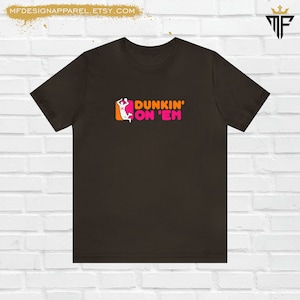 May include: A dark brown t-shirt with the text "DUNKIN' ON 'EM" in pink and orange. The design includes a basketball player silhouette. The Etsy shop name is visible at the top.