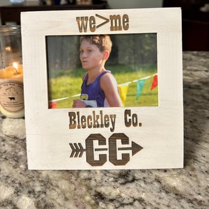 May include: White wooden picture frame with a photo of a young person running in a cross country race. The frame has the text "we>me" at the top, "Bleckley Co." in the middle, and "CC" with arrows on either side at the bottom.