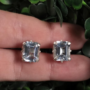 May include: A pair of stud earrings featuring rectangular, emerald-cut light blue gemstones set in silver-toned metal prongs. The stones have a clear, faceted surface that reflects light, and each earring is secured with a four-prong setting.