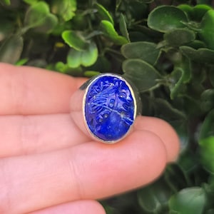 May include: A silver ring featuring a deep blue oval-shaped stone set in the center. The stone is carved with a detailed profile of a figure facing right, showcasing intricate textures and a polished finish on the surrounding silver band.