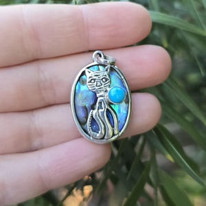 May include: Oval silver pendant featuring a cat design. The cat is set against an iridescent abalone shell background, with a turquoise-colored stone accent. The pendant is held in a hand.