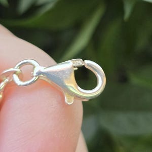 May include: A silver-toned lobster claw clasp attached to a chain link. The metal surface of the clasp features a small, clearly engraved 925 hallmark, indicating sterling silver composition. The clasp mechanism is shown in an open position.