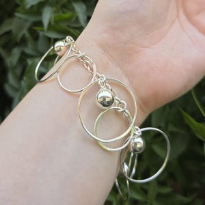 May include: A silver-toned charm bracelet featuring a series of interconnected circular metal rings and polished spherical beads. The bracelet is constructed with a link chain base that supports the dangling circular elements and smooth, reflective spheres.