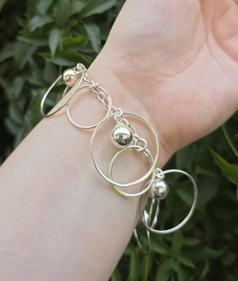May include: A silver-toned charm bracelet featuring a series of interconnected circular metal rings and polished spherical beads. The bracelet is constructed with a link chain base that supports the dangling circular elements and smooth, reflective spheres.