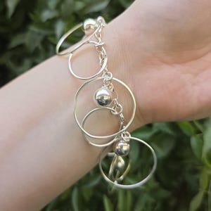 May include: A silver-toned charm bracelet featuring multiple thin, circular metal rings and polished spherical beads attached to a central chain link structure. The bracelet is composed of reflective metallic components arranged in a dangling design.
