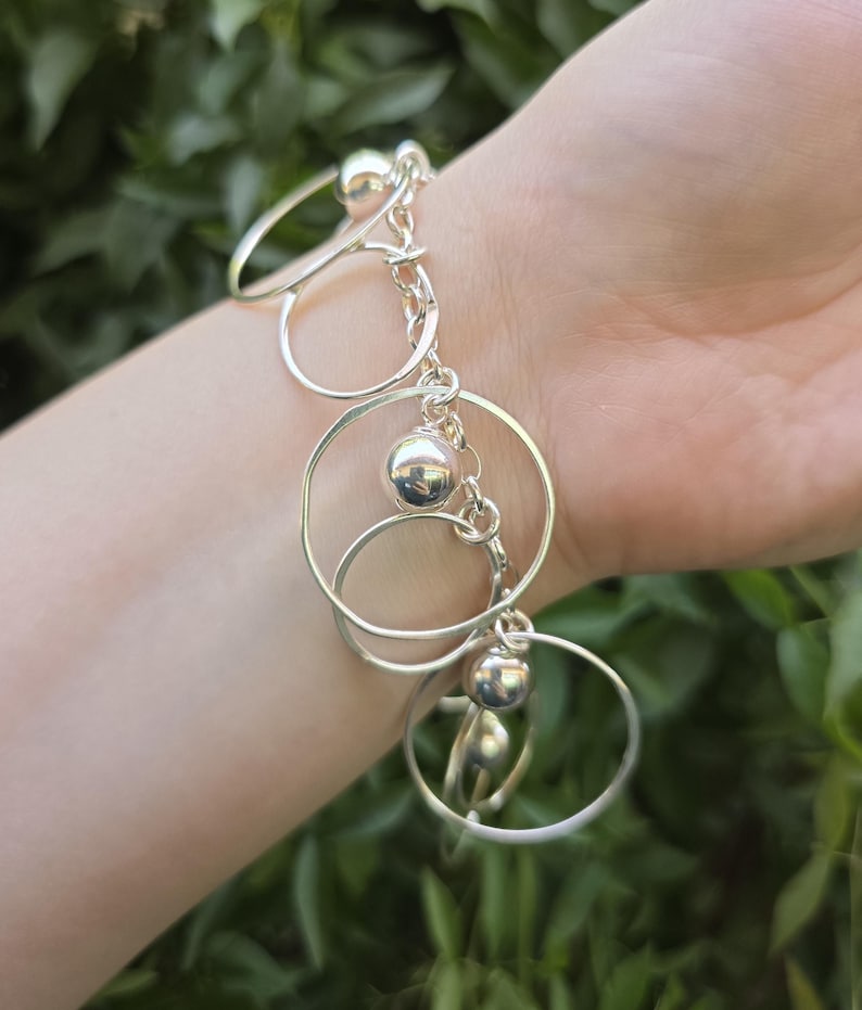 May include: A silver-toned charm bracelet featuring multiple thin, circular metal rings and polished spherical beads attached to a central chain link structure. The bracelet is composed of reflective metallic components arranged in a dangling design.
