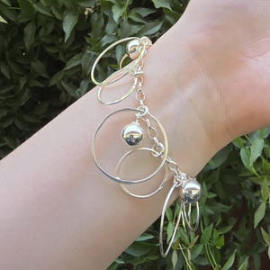 May include: A silver charm bracelet featuring a series of polished metal spheres and multiple open circular rings of varying sizes attached to a linked chain. The bracelet is constructed from reflective silver-toned metal components.
