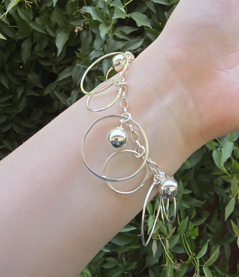 May include: A silver charm bracelet featuring a series of polished metal spheres and multiple open circular rings of varying sizes attached to a linked chain. The bracelet is constructed from reflective silver-toned metal components.