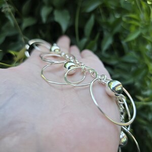 May include: A silver-toned bracelet featuring a series of interconnected thin circular metal rings and polished spherical beads. The design consists of alternating smooth round beads and open wire circles linked together to form a chain-like structure.