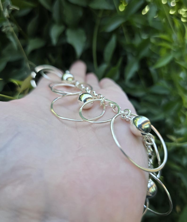 May include: A silver-toned bracelet featuring a series of interconnected thin circular metal rings and polished spherical beads. The design consists of alternating smooth round beads and open wire circles linked together to form a chain-like structure.