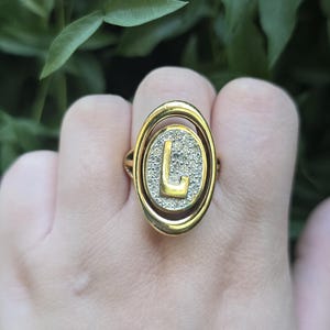 May include: A gold-toned ring featuring an oval-shaped centrepiece with a textured, shimmering surface. A prominent, smooth gold letter L is centred on the oval. The ring has a double-layered oval frame design surrounding the central letter.