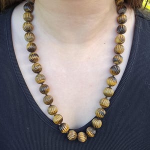 May include: A beaded necklace featuring large, round beads with a ribbed texture. The beads are a mix of golden brown and dark brown, strung together on a dark cord. The necklace is worn against a black top.