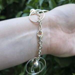 May include: A silver-toned bracelet featuring a series of interconnected circular metal rings and a central lobster claw clasp. The design includes a small, smooth spherical bead charm attached to the chain links, creating a layered metallic look.