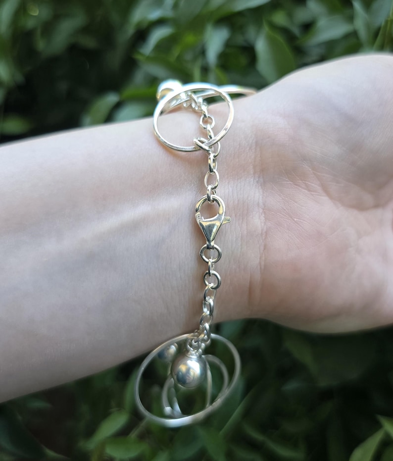 May include: A silver-toned bracelet featuring a series of interconnected circular metal rings and a central lobster claw clasp. The design includes a small, smooth spherical bead charm attached to the chain links, creating a layered metallic look.