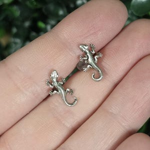 May include: A pair of silver-toned metal stud earrings shaped like small lizards. Each earring features a detailed, textured body with four legs and a curved tail, designed to resemble a gecko or similar reptile in a metallic finish.