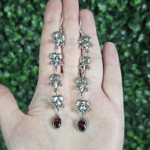 May include: A pair of long silver dangle earrings featuring four stacked floral-shaped metal links connected by small rings, terminating in a single oval-cut deep red garnet gemstone set in a silver bezel at the base of each earring.