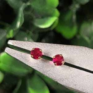 May include: Two small round stud earrings featuring vibrant red faceted stones set in gold-toned metal prongs. The earrings are held securely between the flat metal jaws of a pair of silver-colored pliers, showcasing the circular cut of the gems.