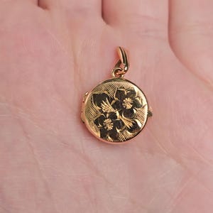 Op de afbeelding: Een goudkleurige ronde locket hanger met een bloemenmotief. De hanger heeft een getextureerd oppervlak en een kleine lus voor een ketting. Het bloemenmotief is in het metaal geëtst.