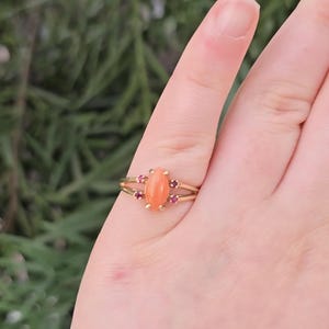 May include: A gold ring featuring an oval, peach-colored gemstone, flanked by smaller, round, red gemstones. The ring has a split band design. The ring is displayed on a finger against a blurred green background.