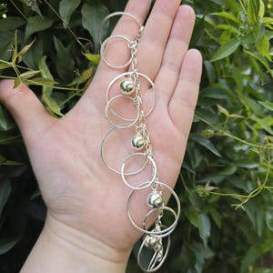 May include: A silver-toned charm bracelet featuring a series of interconnected circular rings of varying sizes and polished spherical beads attached to a central chain link structure, creating a layered and dangling metallic design.