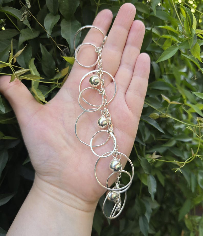 May include: A silver-toned charm bracelet featuring a series of interconnected circular rings of varying sizes and polished spherical beads attached to a central chain link structure, creating a layered and dangling metallic design.