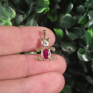 May include: A gold-toned pendant shaped like a teddy bear, featuring a clear round gemstone for the head and an oval-shaped deep pink gemstone for the body. The metal limbs and ears have a textured finish, and a small bail is attached at the top.