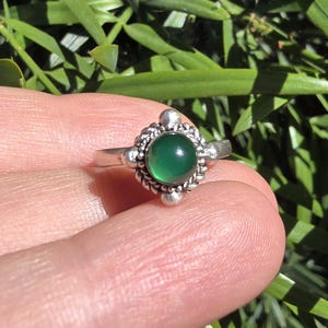 May include: A silver ring with a round, vibrant green gemstone at the center. The gemstone is surrounded by a detailed, ornate silver setting. The ring is held against a backdrop of green foliage.