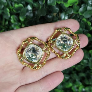 May include: A pair of gold-tone earrings featuring a square-shaped clear faceted crystal set in the center. The crystal is surrounded by a decorative frame made of twisted gold-tone metal wire that forms a square border with rounded corners.