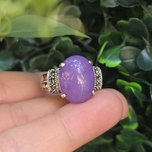 May include: A ring featuring a large, smooth, oval-shaped purple stone set in a silver band. The setting includes small, dark, faceted stones arranged in a decorative pattern on both sides of the central purple cabochon, secured by four prongs.