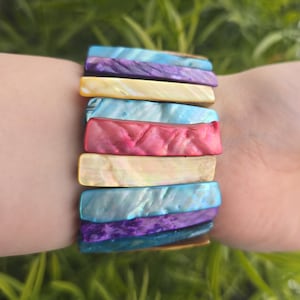 Könnte beinhalten: Ein farbenfrohes Armband mit rechteckigen, irisierenden Segmenten in Blau-, Lila-, Rosa- und Gelbtönen. Das Armband besteht aus natürlichen Materialien und wird am Handgelenk getragen. Die Segmente des Armbands sind etwa 2,5 cm lang.