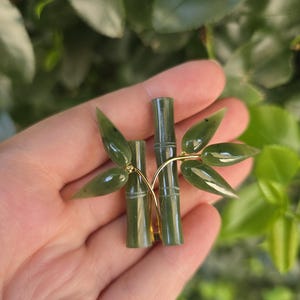 Puede incluir: Un broche de bambú de jade verde con detalles dorados. El broche presenta dos tallos de bambú y varias hojas de jade. Las hojas y los tallos son de color verde oscuro. El broche se sostiene en una mano sobre un fondo de follaje verde.