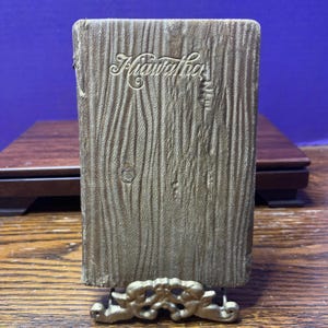 Puede incluir: Un libro antiguo con el título "Hiawatha" en relieve en una escritura ornamentada. La cubierta tiene un aspecto texturizado de vetas de madera en color marrón claro. El libro está apoyado en un soporte decorativo dorado.