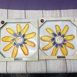 Puede incluir: Dos obras de arte enmarcadas, cada una con una gran flor amarilla con detalles azules y naranjas. Los marcos cuadrados son de color claro. La marca "olika" es visible en la esquina superior izquierda de cada pieza. El texto "INTERCONTINENTAL ART INCORPORATED" es visible.