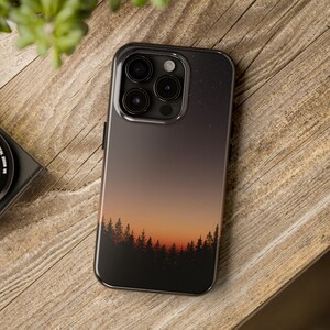 May include: A black phone case with a gradient design featuring a sunset over a forest silhouette. The case is shown on an iPhone 15 Pro.