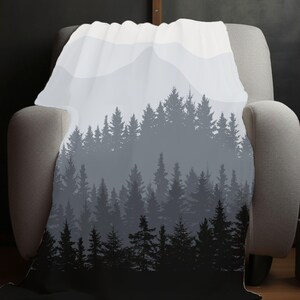 May include: A soft, white throw blanket with a black and grey silhouette of a forest and mountains. The blanket is draped over a grey armchair.