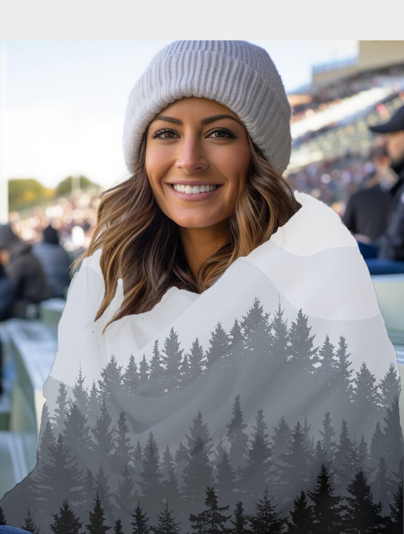 May include: A woman wearing a white knitted beanie and a grey and white scarf with a forest silhouette print. The scarf is draped over her shoulders and the forest silhouette is visible in the background.