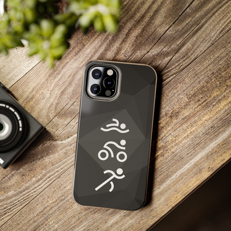 May include: A dark gray smartphone case featuring a geometric low-poly pattern and a white graphic depicting a triathlon sequence with a swimmer, a cyclist, and a runner stacked vertically in the center of the back panel.