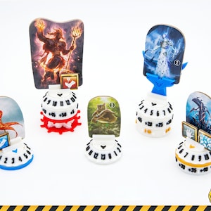 Puede incluir: Cinco contadores de juego circulares de plástico blanco con diales numerados y bases de colores. Cada contador sostiene una tarjeta de personaje ilustrada vertical con figuras de fantasía, incluyendo un guerrero de fuego y un ser de hielo.