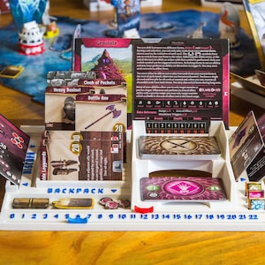 Pode incluir: Um organizador de jogos de tabuleiro em pl&aacute;stico branco com ranhuras para cartas, uma pista numerada de 0 a 22 e etiquetas incluindo Backpack e Discard. O organizador cont&eacute;m v&aacute;rias cartas de jogo com ilustra&ccedil;&otilde;es de um machado de batalha e grevas.