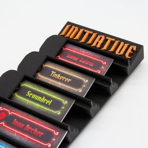 May include: A black plastic tabletop game organizer featuring the word INITIATIVE in orange lettering at the top. It contains four rectangular character cards labeled Stone Golem, Tinkerer, Scoundrel, and Inox Archer, each with distinct colored borders.