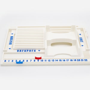 Pode incluir: Um organizador de jogos de tabuleiro em pl&aacute;stico branco com uma faixa numerada de 0 a 22 em texto azul. Inclui sec&ccedil;&otilde;es designadas com as etiquetas DISCARD, BACKPACK e LOST em azul, com um marcador deslizante azul e um vermelho.