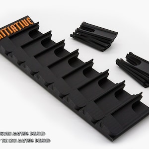 May include: A black plastic initiative tracker with orange text reading INITIATIVE, featuring eight slots for game cards. Included are two separate sets of eight black plastic adapters, labeled as Frosthaven and Jaws of the Lion adapters.