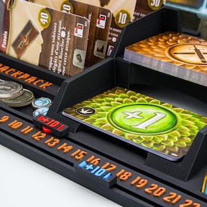 Pode incluir: Um organizador de jogos de tabuleiro preto impresso em 3D com uma se&ccedil;&atilde;o de mochila, compartimentos para cartas e uma trilha de contagem integrada com n&uacute;meros laranja de 6 a 22. Cont&eacute;m v&aacute;rias cartas de jogo, incluindo uma com um s&iacute;mbolo verde de mais um.
