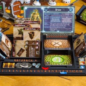 Pode incluir: Um organizador de jogos de tabuleiro em pl&aacute;stico preto com um painel de personagem Brute, v&aacute;rias ranhuras para cartas com texto em franc&ecirc;s, uma &aacute;rea de armazenamento de mochila e uma pista numerada de 0 a 22 com marcadores.