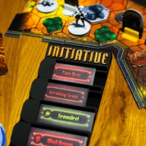 May include: A black plastic board game initiative tracker with the word INITIATIVE in gold lettering. It holds four rectangular cards labeled Cave Bear, Vermling Scout, Scoundrel, and Wind Demon, positioned next to a hexagonal game map with miniatures.