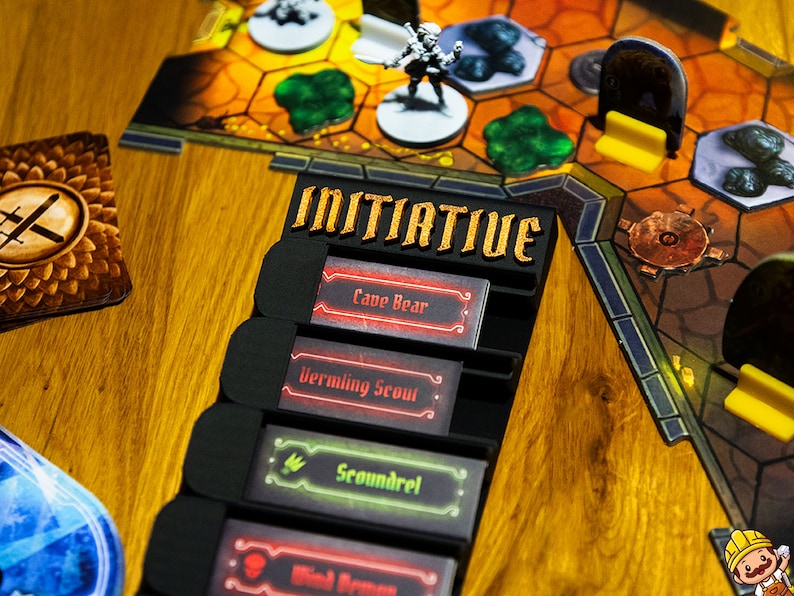 May include: A black plastic board game initiative tracker with the word INITIATIVE in gold lettering. It holds four rectangular cards labeled Cave Bear, Vermling Scout, Scoundrel, and Wind Demon, positioned next to a hexagonal game map with miniatures.