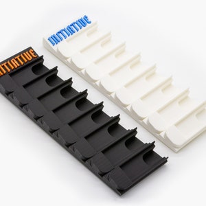 May include: Two rectangular game tracker organizers, one black and one white, each featuring seven slots for game pieces. The black organizer has the word INITIATIVE embossed in orange, while the white organizer has the same word in blue.