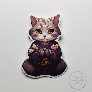 Può includere: Un gatto grigio e bianco che indossa una veste nera con fiori rosa. Il gatto è seduto con le zampe incrociate davanti a sé.