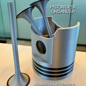 May include: A silver-colored 3D-printed piston desk organizer with three black-accented valve stem pens. The organizer features a cylindrical body with horizontal black rings and internal compartments. Text on the image reads PISTON DESK ORGANIZER and INCLUDES 3 VALVE STEM PENS.